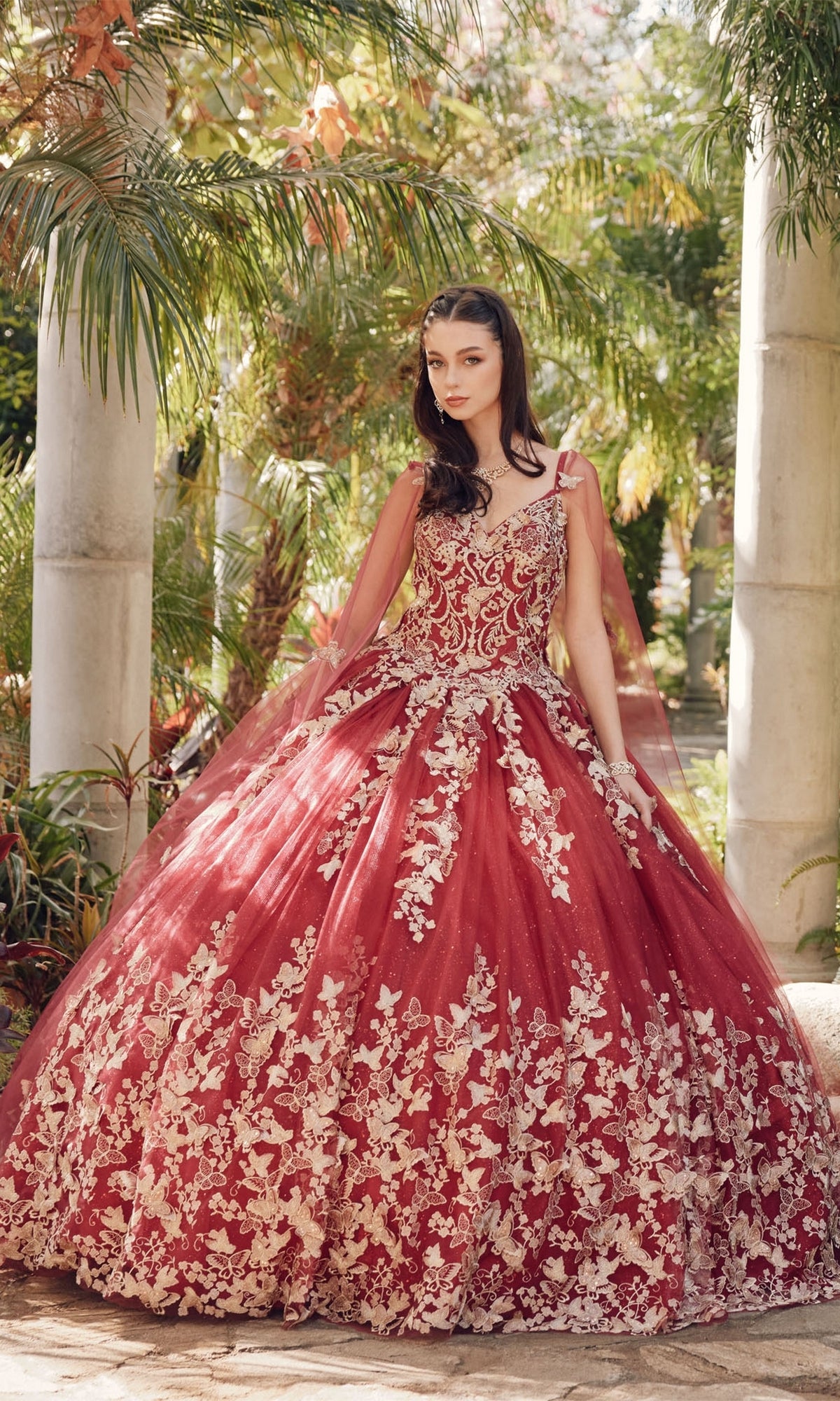 A woman stands outdoors in the Juliet 1442 Quinceañera Dress with Sheer Cape, a voluminous burgundy gown featuring intricate gold floral embroidery, surrounded by lush greenery and elegant stone columns.